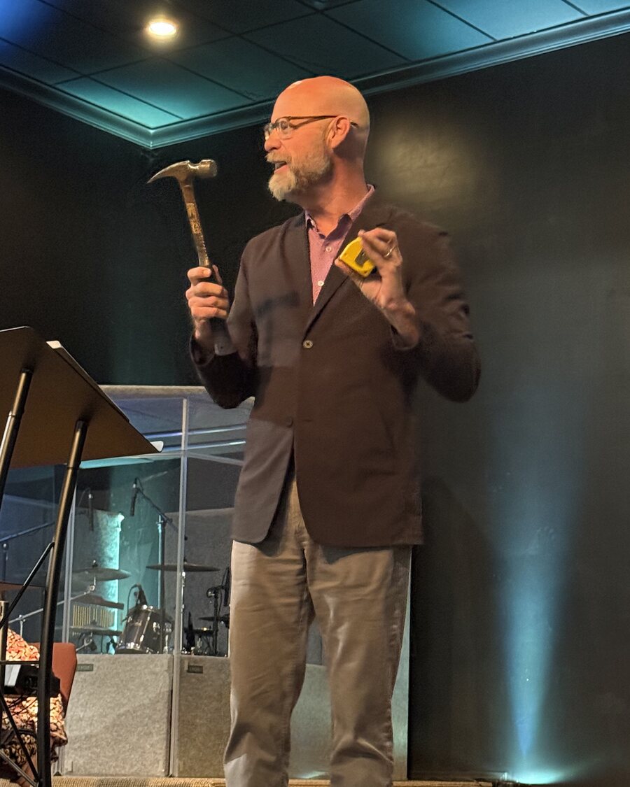 Joe Cook preaching with a hammer and a tape measure in hand
